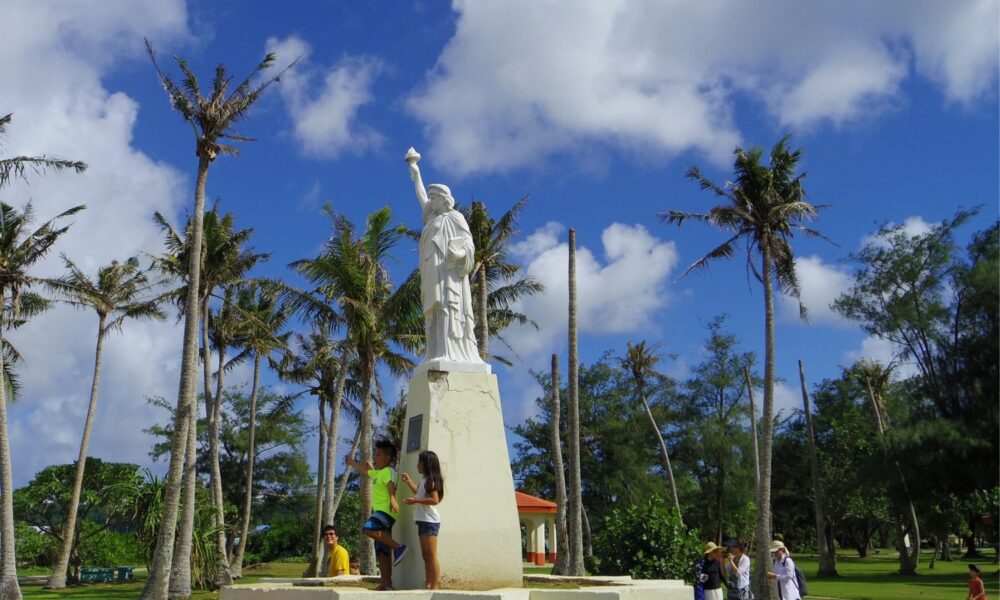 在一个阳光明媚的日子里，在关岛的首都Hagatña的Paseo公园里，几个孩子站在一个按比例缩小的自由女神像复制品前。