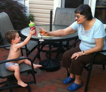 一位年长的妇女和一个婴儿坐在院子里的桌子旁。女人端着一杯酒，向婴儿敬酒，婴儿也用吸管杯敬酒。