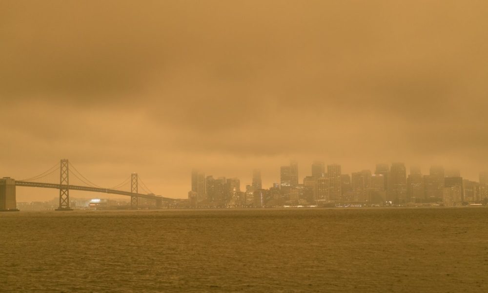 Haze from wildfire smoke obscures a view of San Francisco from across the water. Everything in the photo appears yellow, orange, or brown with the pollution in the air.