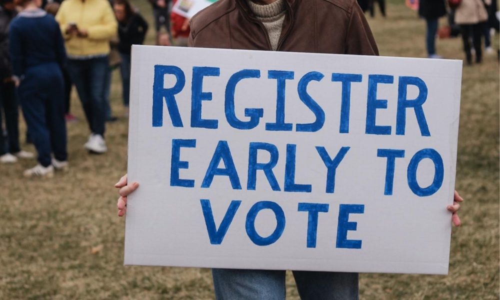 一名身穿棕色皮夹克的白人男子举着一块牌子，上面写着“提前登记投票”。