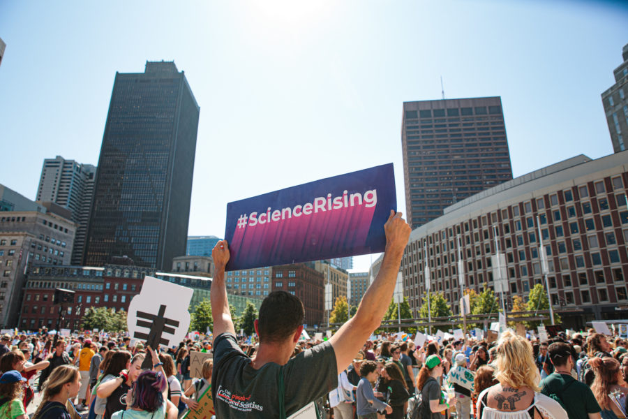 在一个拥挤的广场上，一名男子举着紫色标语，上面写着#ScienceRising，他和其他许多人站在一起，其中一些人也举着标语。背景是摩天大楼。