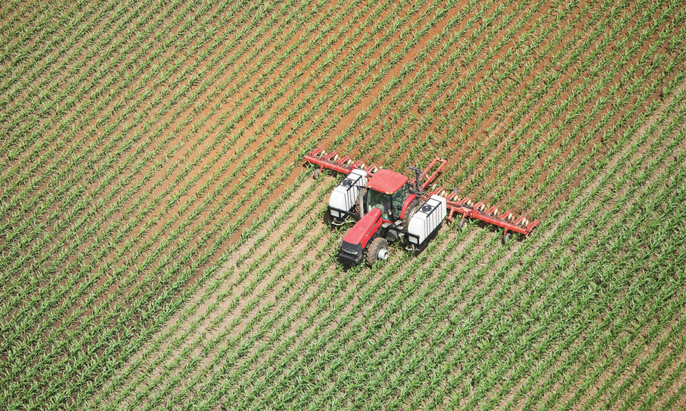 鸟瞰图，一辆拖着施药机的拖拉机正在晚春的玉米地里喷洒液氮肥料。左边的田地和撒播机后面的田地被深色的土壤弄湿了。从一架小飞机开着的窗户里射出的。