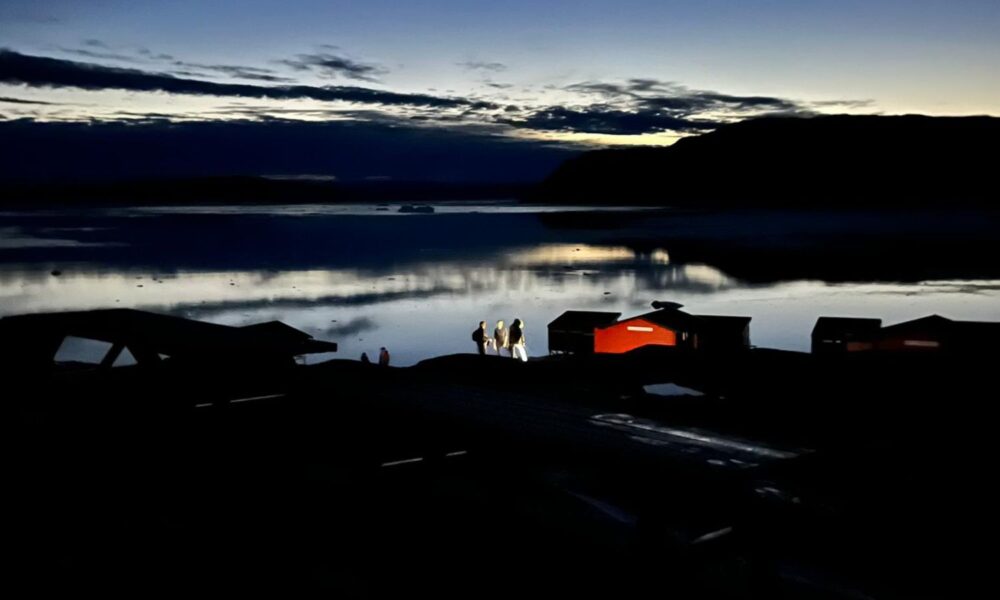 在暮色中，从远处看，三个女人拿着手电筒站在一间小屋的入口处。背景中可以看到一个湖。