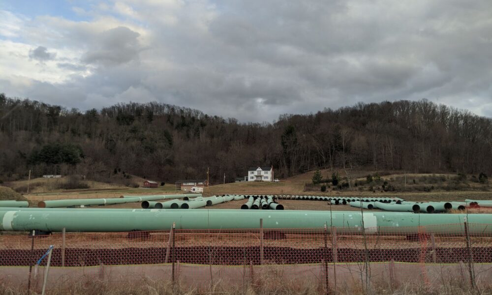 在前景中，一系列用于输送天然气的绿色管道坐落在弗吉尼亚州或西弗吉尼亚州的丘陵景观上，背景中可以看到一座房子
