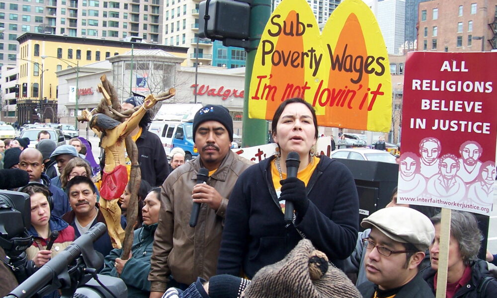 这张照片是一名女子在城市街道上通过麦克风对人群讲话，她面前的牌子上写着“低于贫困线的工资:我不喜欢它”。