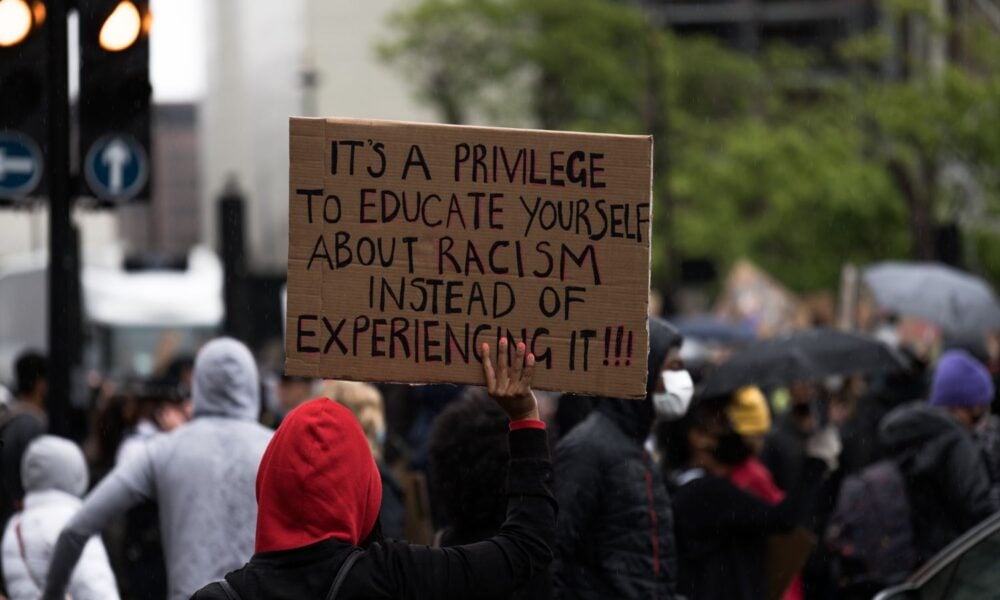 在一场抗议活动中，一个人的手举着一个牌子，上面写着:“教育自己种族主义是一种特权，而不是经历种族主义!!”