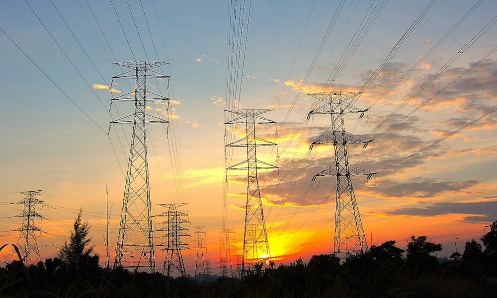 photo of several large towers supporting electricity transmission lines that stretch high above the camera, with a bright orange sunset in the background