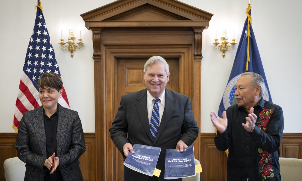 美国农业部长汤姆·维尔萨克微笑着拿着美国农业部公平委员会2023年中期报告的复印件;他身旁是委员会的一男一女成员，他们都在鼓掌;背景是美国国旗和美国农业部的旗帜
