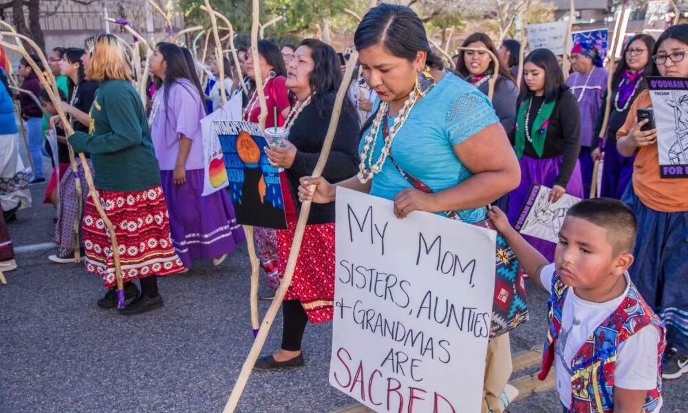 托霍诺印第安妇女领导了2019年图森妇女游行，展示了力量、韧性和权力。这个女人的牌子上写着:我的妈妈、姐妹、阿姨和奶奶都是神圣的。她的儿子在她身边。国际妇女节