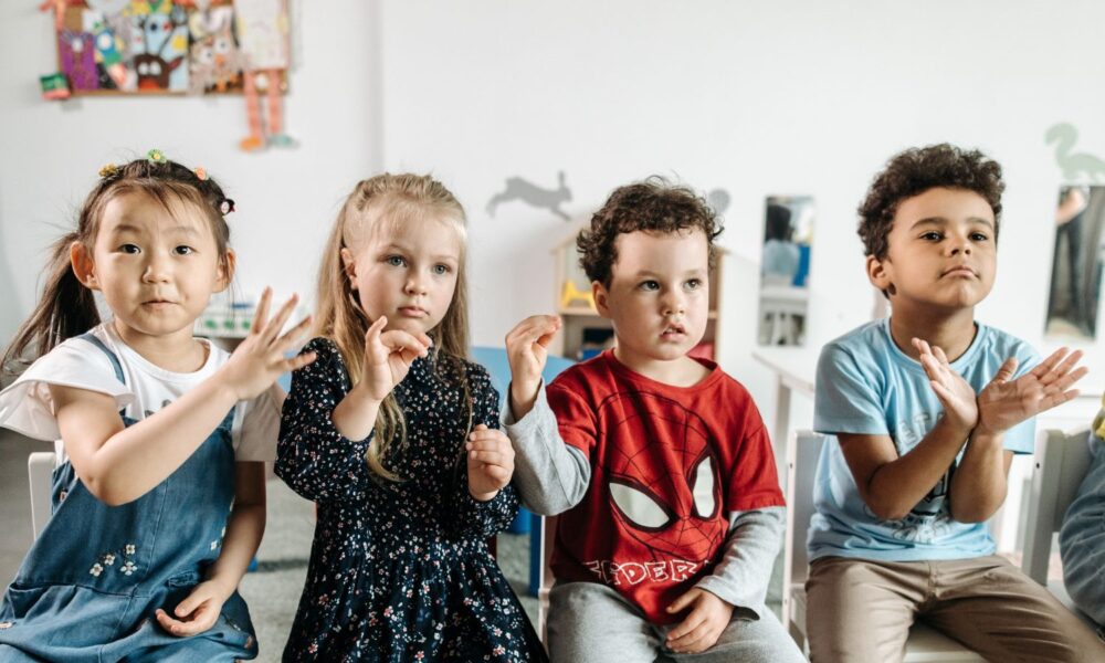 四个不同种族的学龄前儿童坐在一所学校里，举起他们的手，好像他们在背诵一首歌或一节课