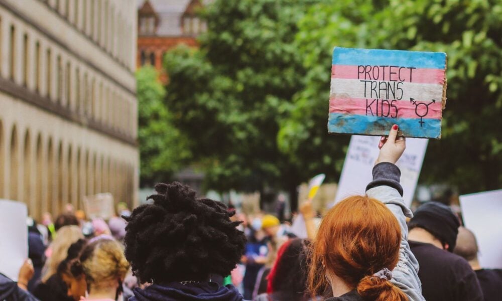 在集会或抗议活动中，有两个人背对着镜头。其中一人举着一个涂有变性人骄傲颜色的牌子，上面写着“保护变性孩子”。