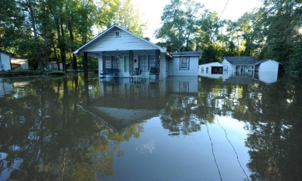 Flood waters surround a modest-looking white house with a detached garage behind it. The water has submerged what you could assume is a driveway, a front porch or steps, and the front and side yards.