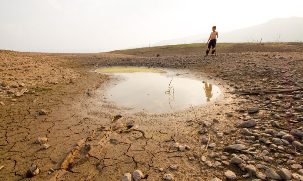 A shirtless kid who looks to be about 10 or 12 years old stands near a small puddle of water that clearly used to be a lake or river, with dry cracked earth all around it. It looks very hot and dry where he is.