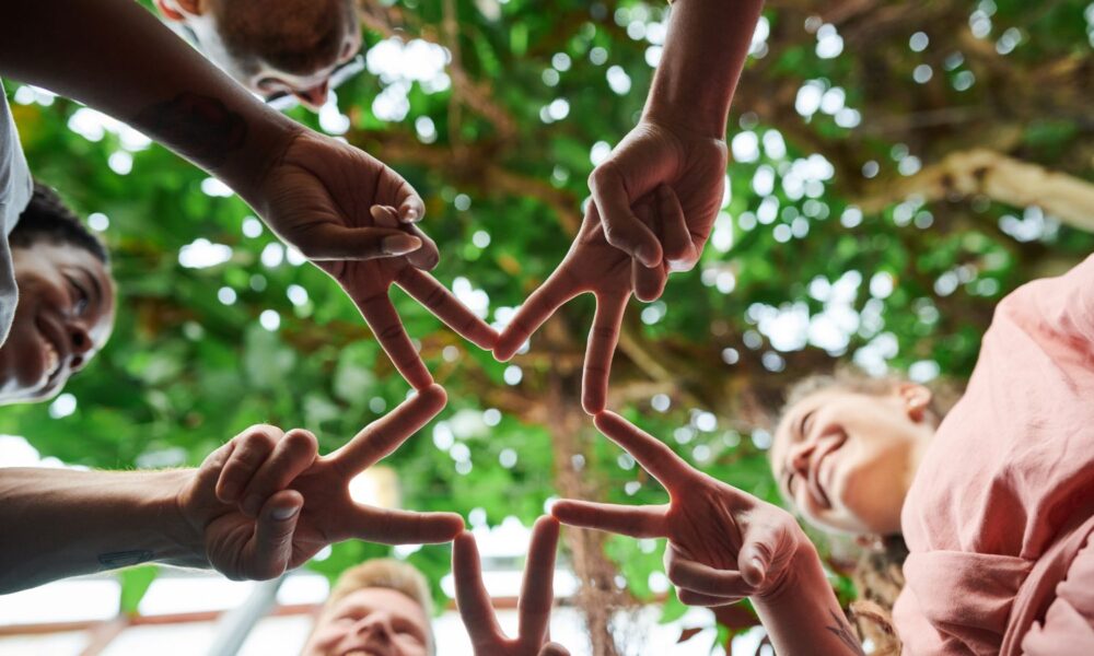 from below, a five-pointed star shape is made up from a diverse group of five people extending two fingers each. the image suggests unity.